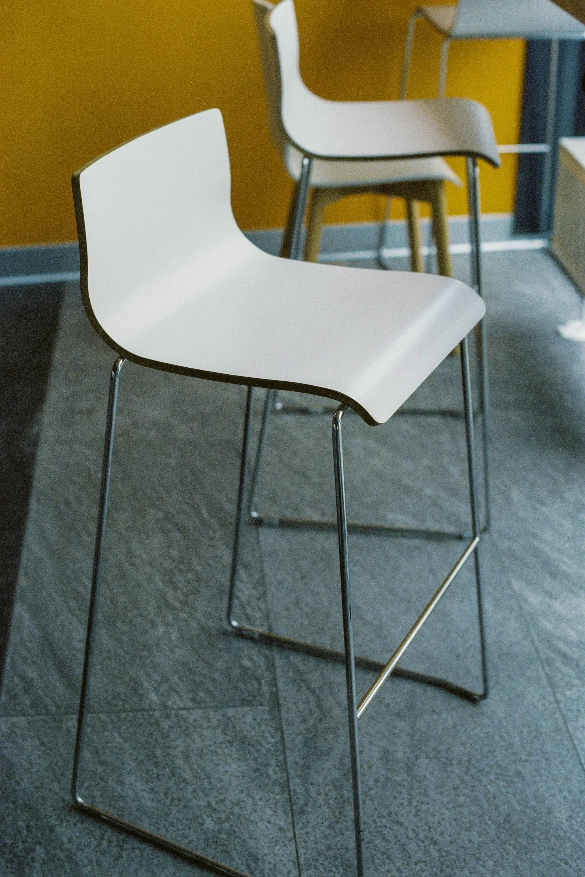 Detail moderní bílé barové židle s ergonomicky tvarovaným skořepinovým sedákem a subtilní chromovanou podnoží typu lyžiny.