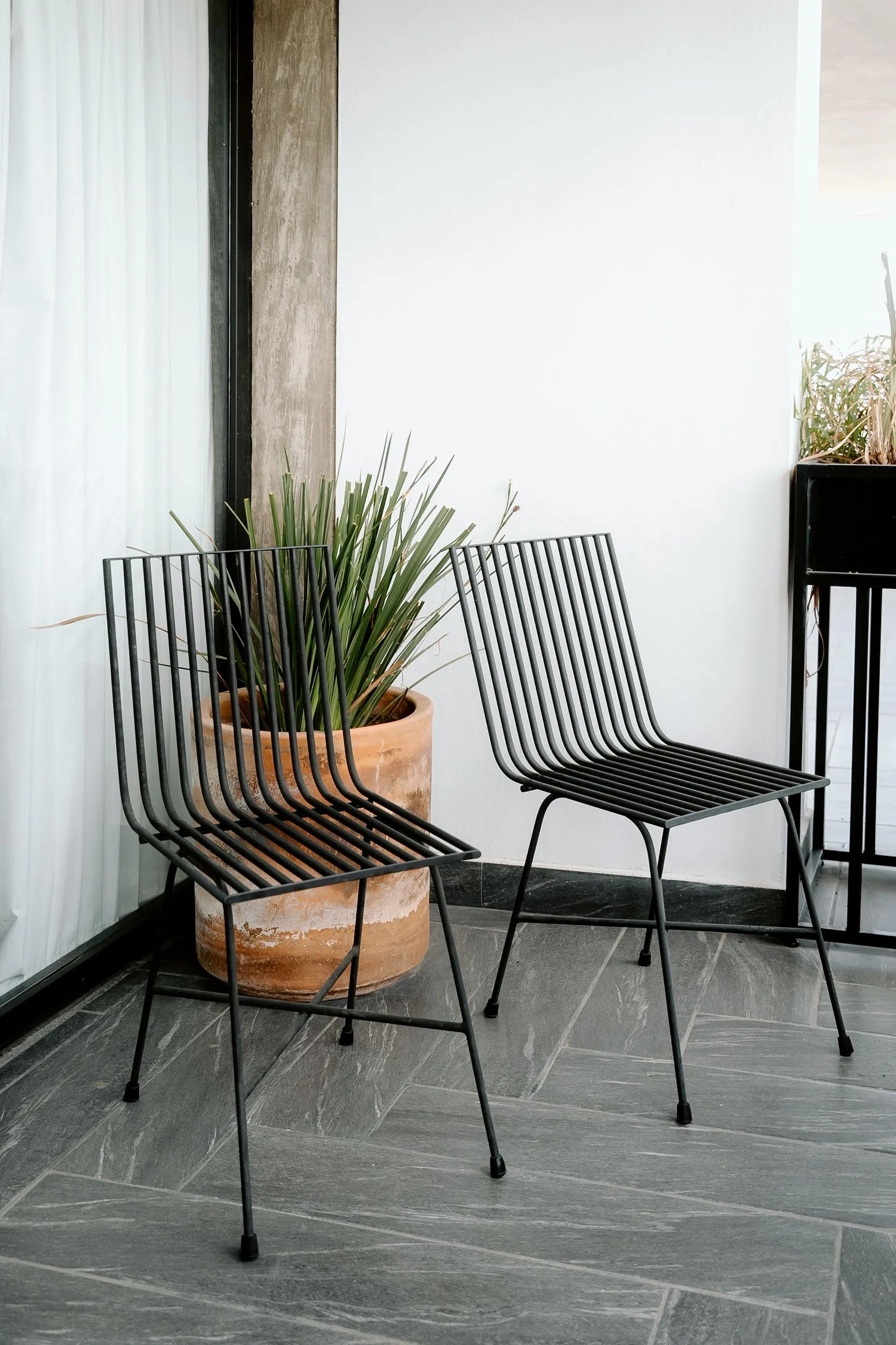 Dvě černé designové židle vyrobené z ohýbaných kovových prutů, umístěné na venkovní terase s kamennou dlažbou a dekorativní rostlinou.
