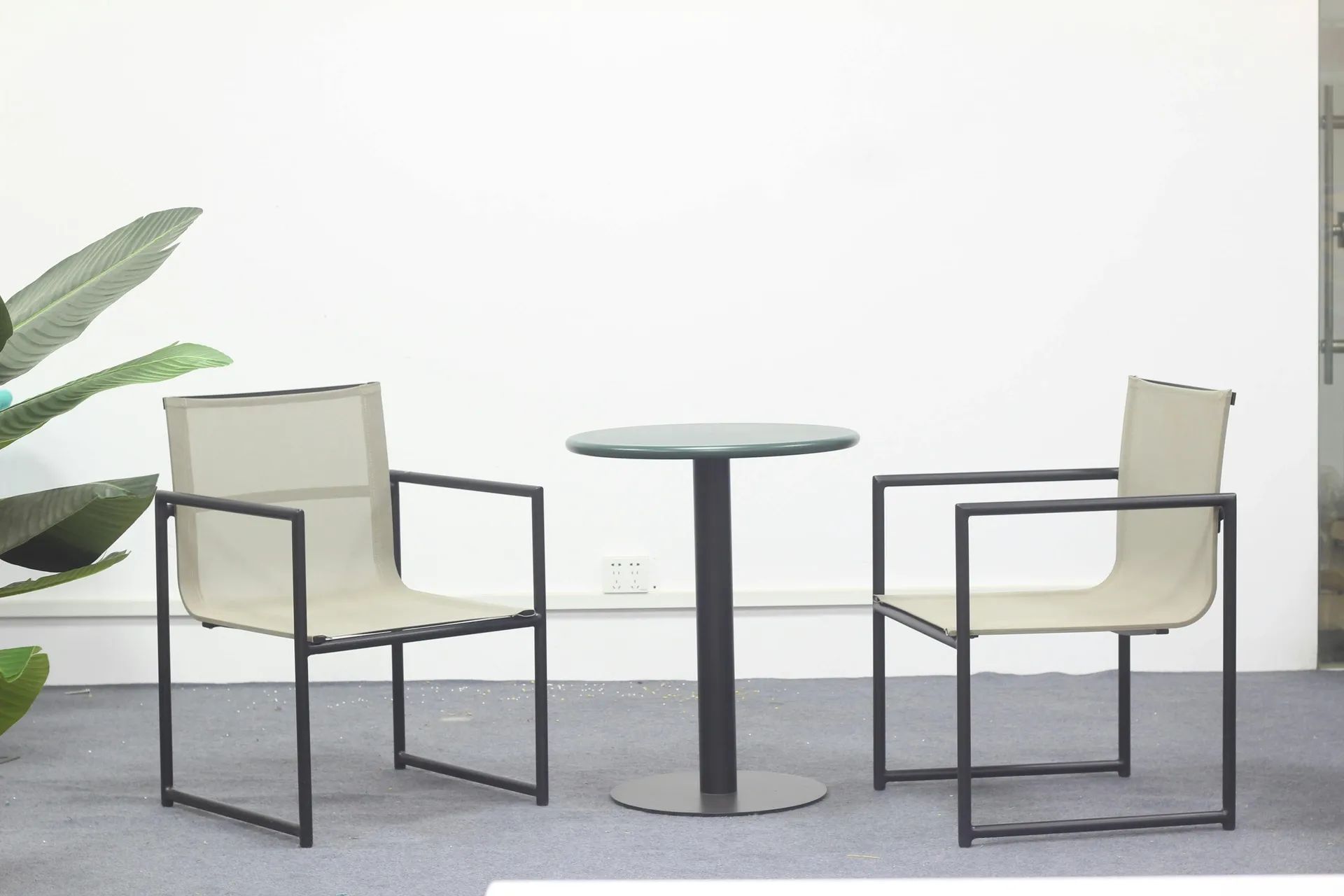 Set dvou minimalistických křesel s černým kovovým rámem a béžovým výpletem, doplněný malým kulatým stolkem na centrální podnoži v čistém bílém prostoru.