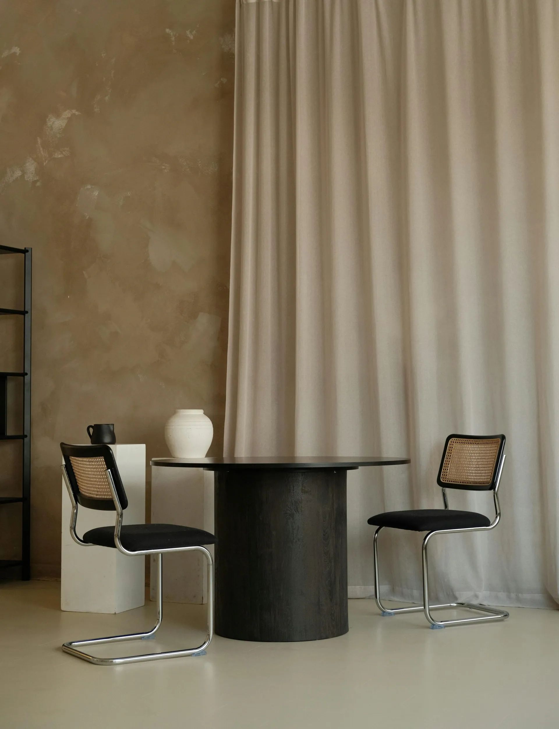 Elegantní jídelní kout s masivním tmavým kulatým stolem a dvěma židlemi v retro stylu, kombinující chromovou konstrukci, černé čalounění a rákosový výplet opěradla.