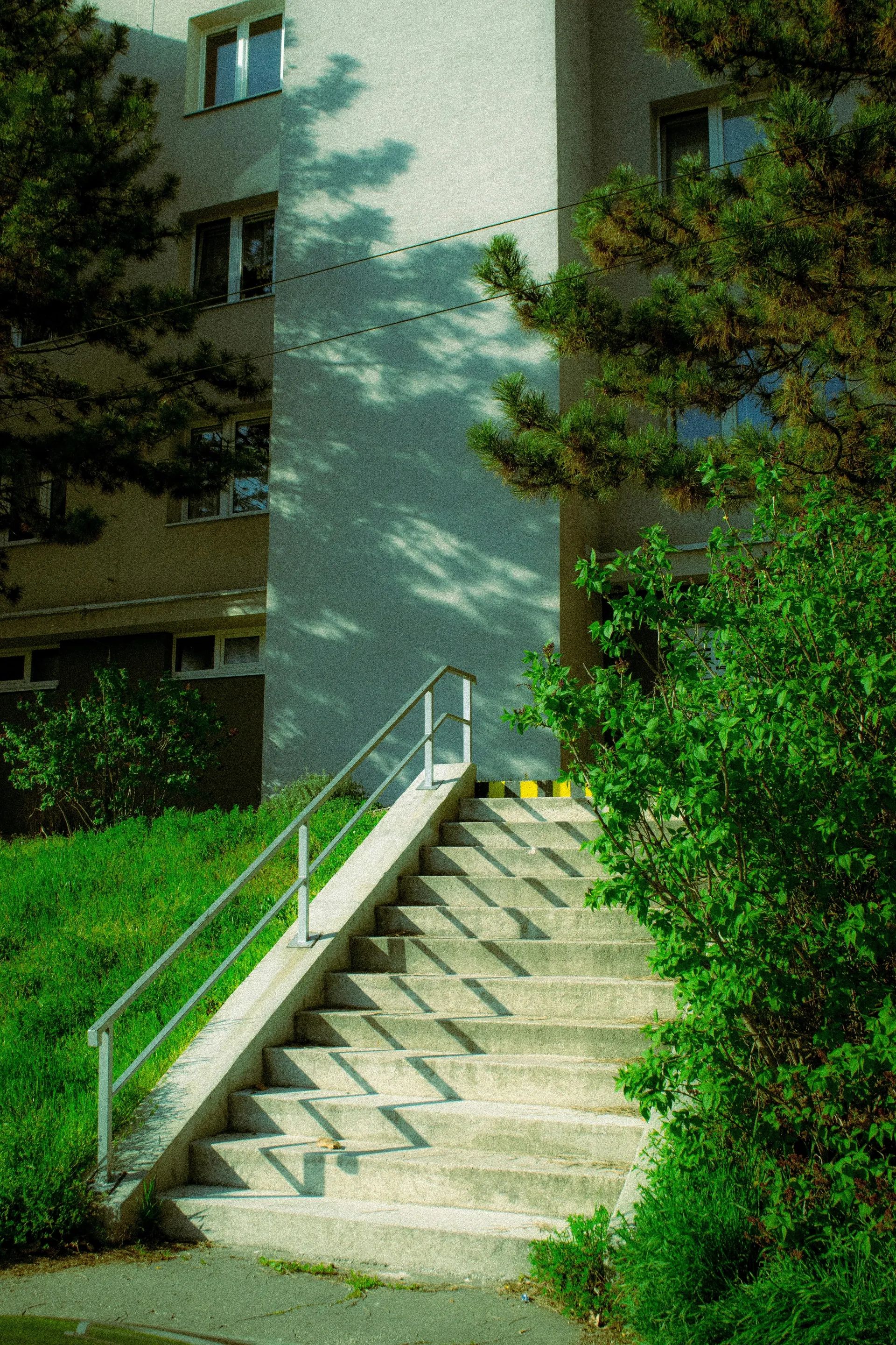 Venkovní betonové schodiště s kovovým zábradlím u bytového domu, obklopené udržovaným trávníkem a zelení za denního světla.