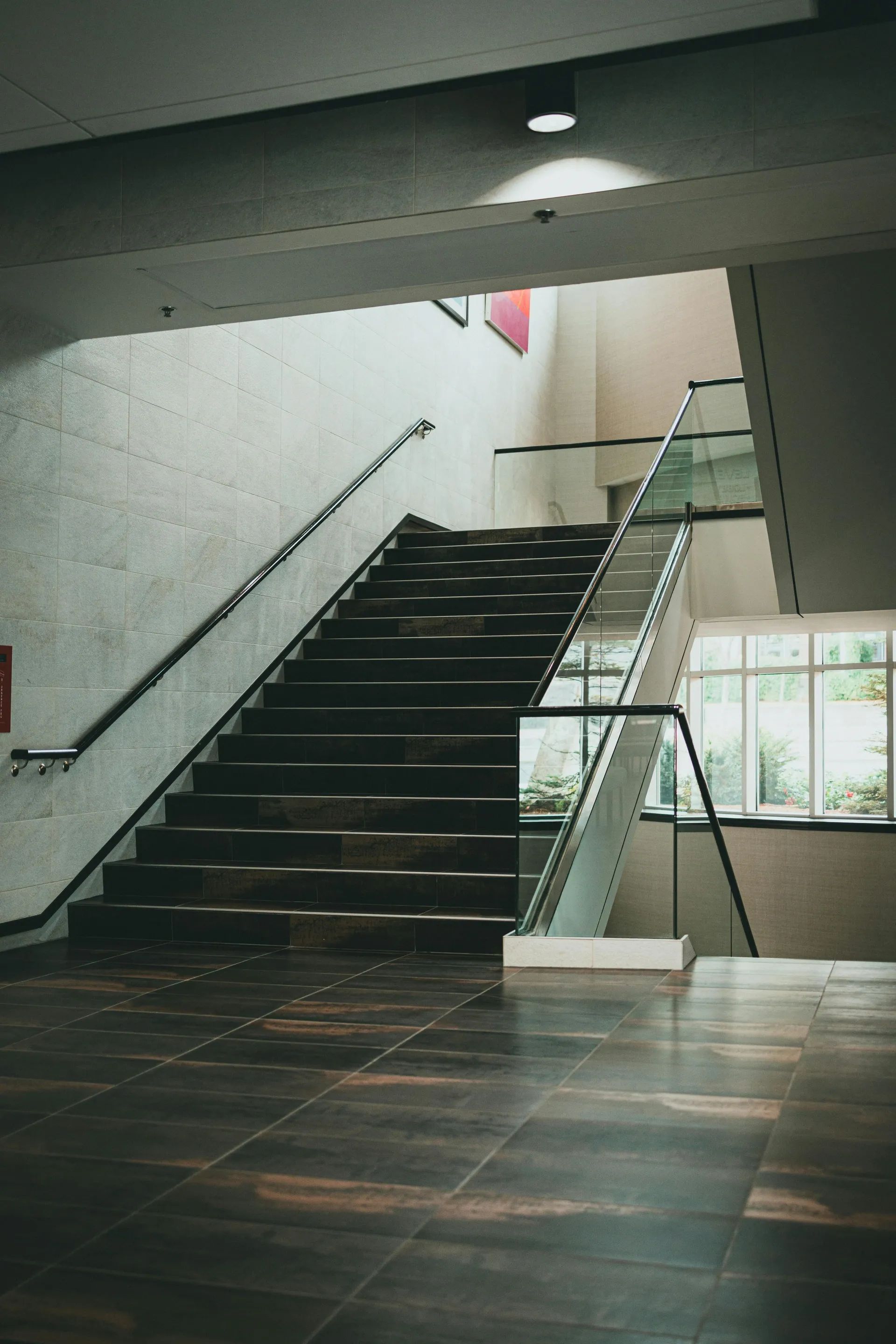 Reprezentativní široké schodiště s tmavým obkladem a moderním celoskleněným zábradlím v osvětleném komerčním interiéru.